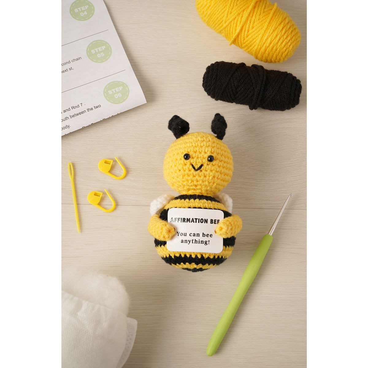 A crocheted bee amigurumi toy sits on a light wood surface with yarn balls, a crochet hook, stitch markers, and a printed instruction sheet. The bee holds a sign that reads, 'I can bee anything!'.