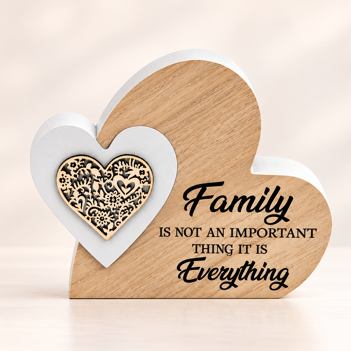 A heart-shaped wooden plaque with text reading 'Family IS NOT AN IMPORTANT THING IT IS Everything' in black script and block lettering. The plaque has a natural light wood finish and is shown with two smaller heart cutouts in front: one white heart and one smaller natural wood heart with an ornate floral pattern.
