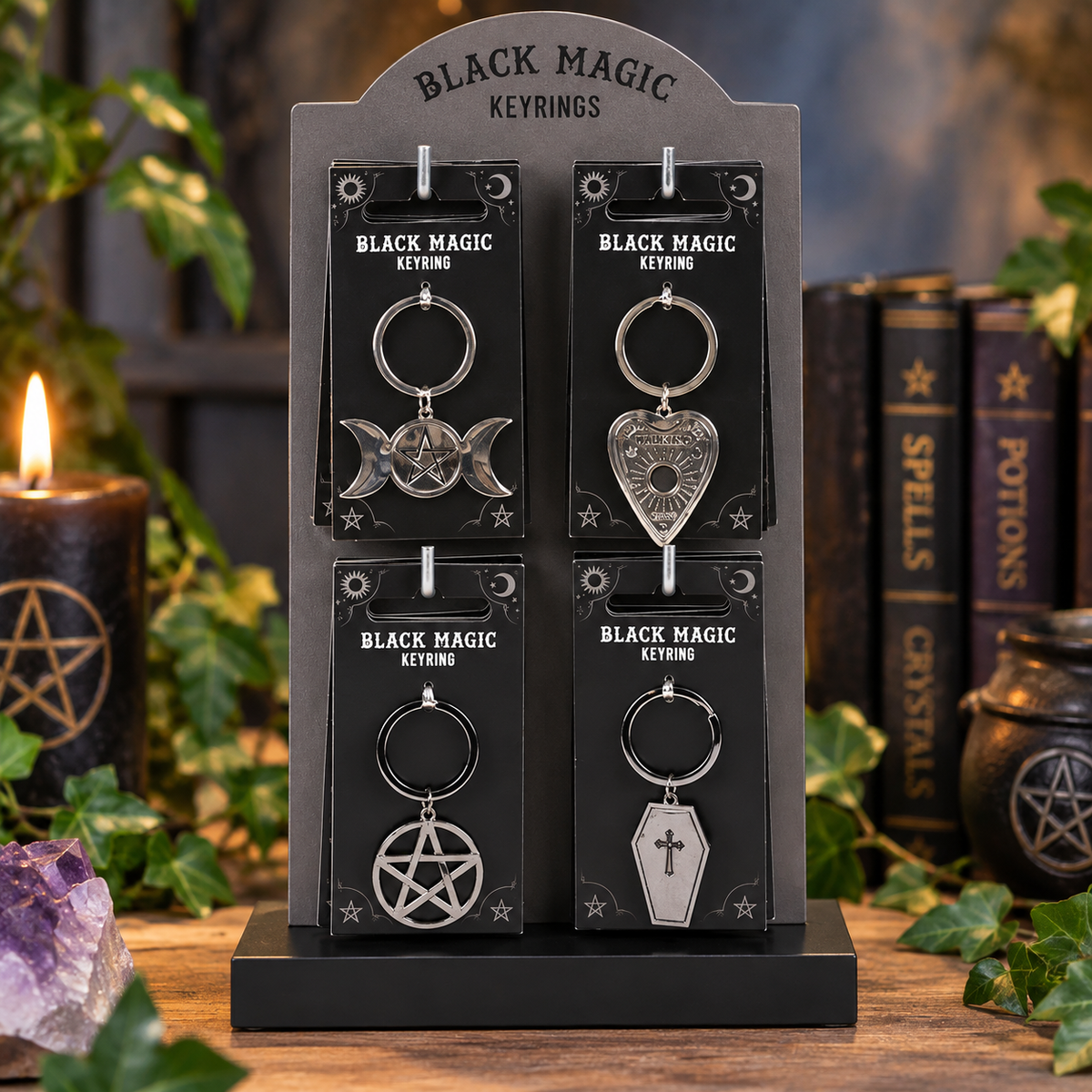 Four black magic keyrings displayed on a black stand, surrounded by themed props including candles, books, and greenery.