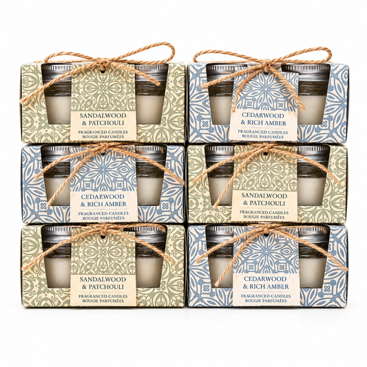 Six boxed scented candles arranged in a 3-by-2 grid on a white background. Each package contains two small glass jar candles tied with twine and a front label reading either Sandalwood & Patchouli or Cedarwood & Rich Amber.