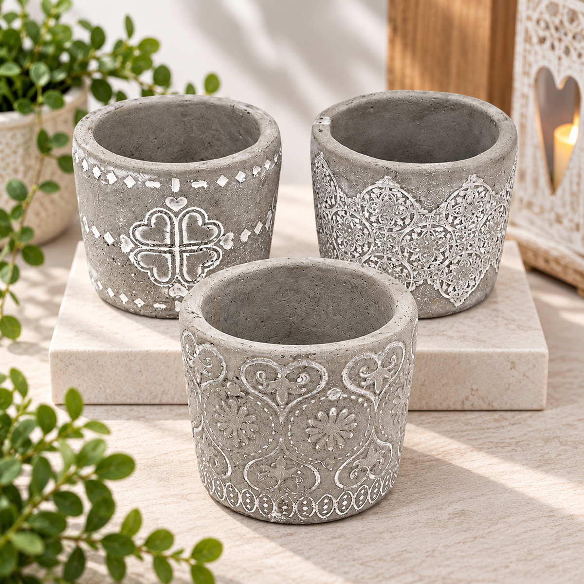 Three small gray decorative planters with embossed floral and geometric patterns, arranged on a light stone block with greenery and a warm lantern in the background.
