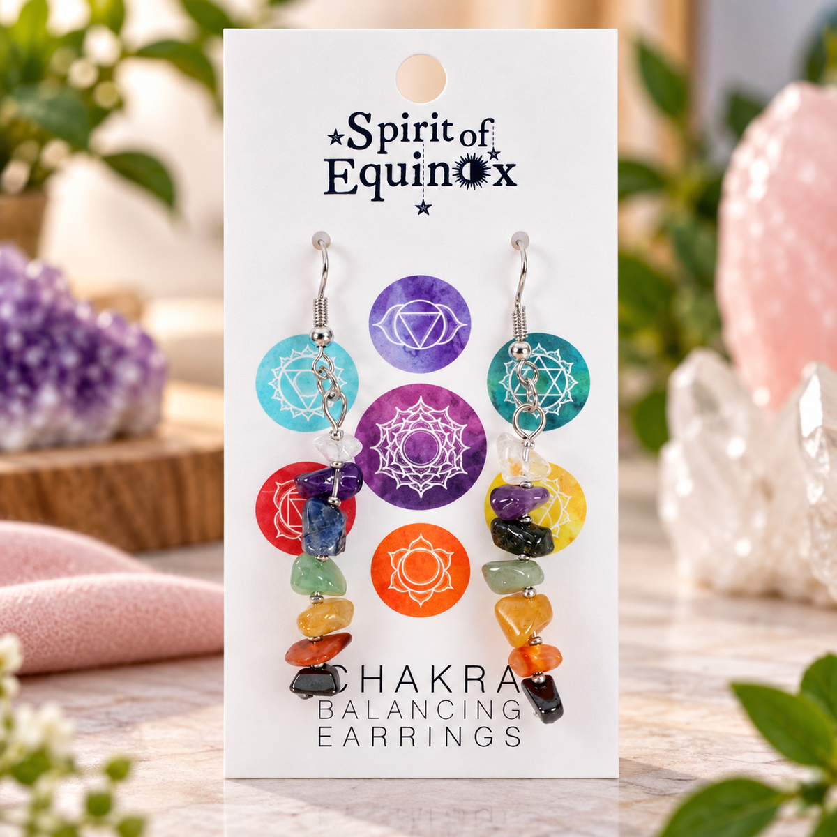 A pair of chakra balancing earrings displayed on a white card labeled 'Spirit of Equinox' and 'Chakra Balancing Earrings.' The earrings feature colorful gemstone chips in red, orange, yellow, green, blue, and purple with white chakra symbols on circular charms.