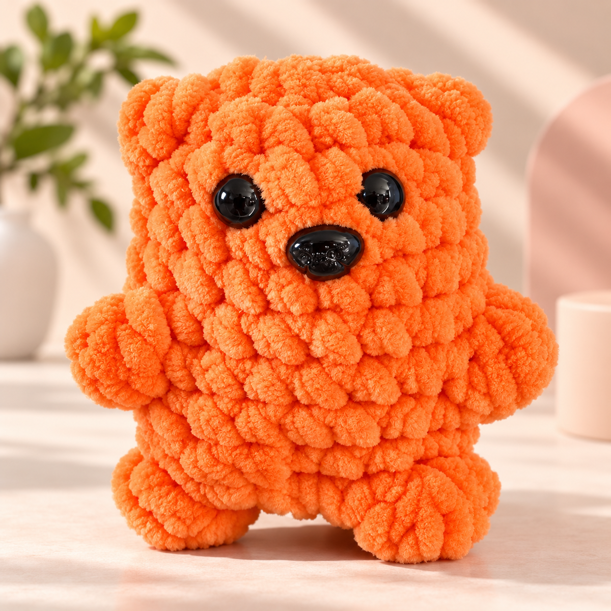 An orange chunky yarn teddy bear toy with black plastic eyes and nose, standing on a light-colored surface with a softly blurred background.