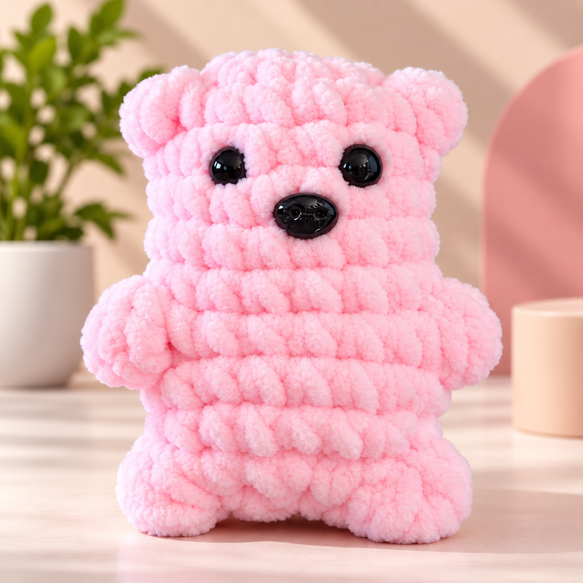 A pink chunky crocheted bear toy with black bead eyes and a black nose, standing on a light surface with a blurred indoor background.