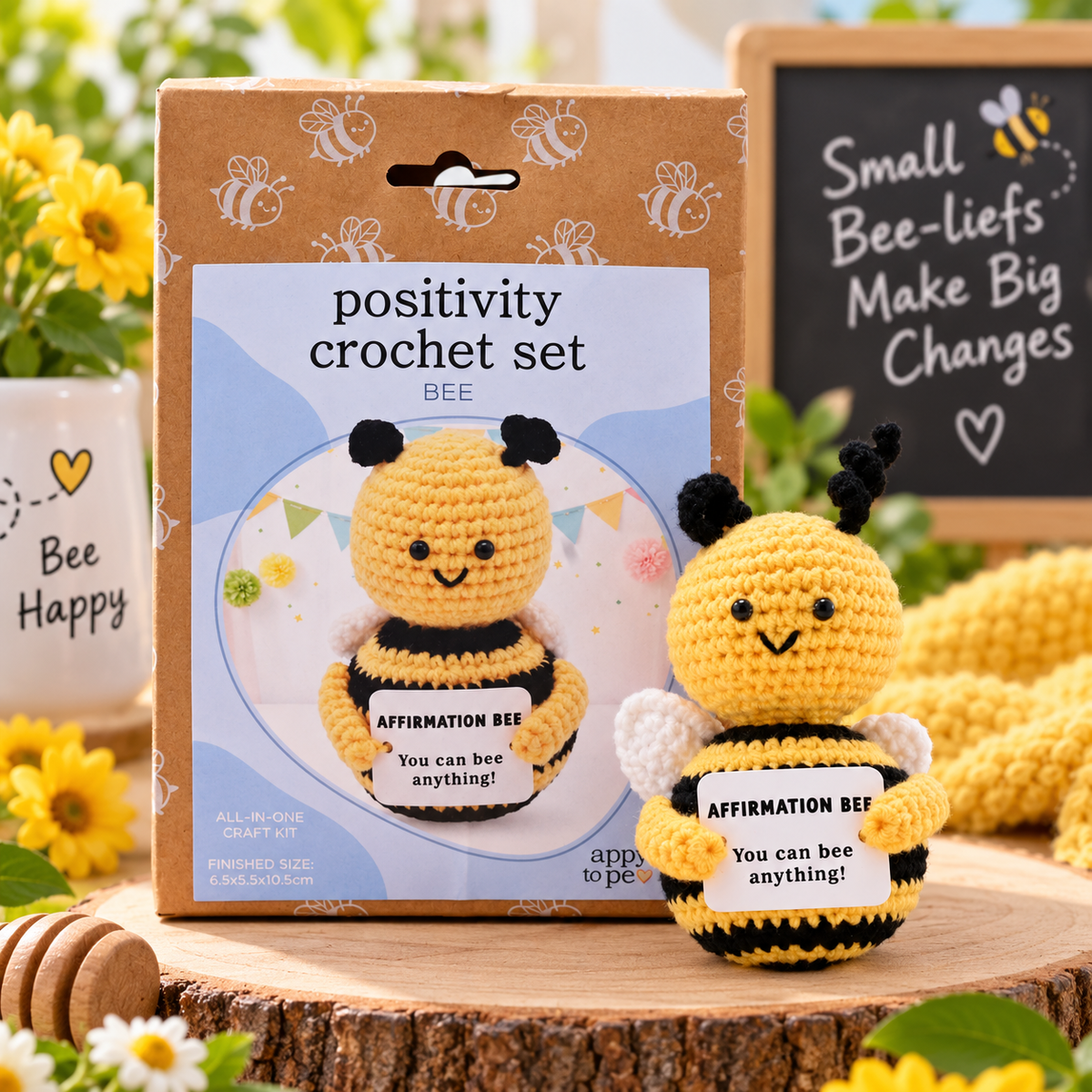 Positivity crochet set 'BEE' kit showing the kraft paper packaging box next to the finished crocheted bee holding a sign that says 'AFFIRMATION BEE: You can bee anything!'. The scene is styled with yellow flowers, a wooden honey dipper, a mug that says 'Bee Happy', and a small chalkboard sign in the background.