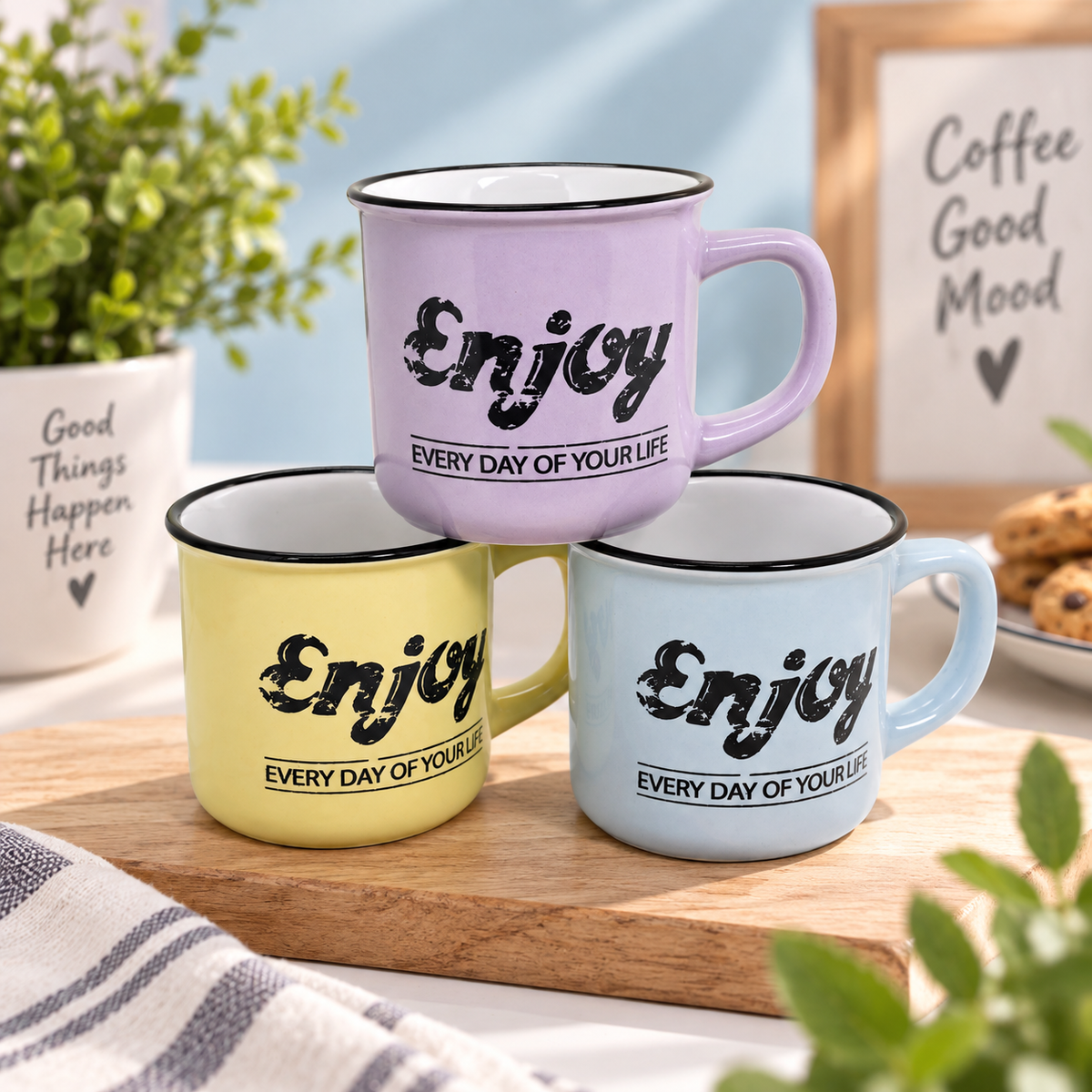 Three enamel-style mugs in pastel purple, yellow, and light blue are stacked on a wooden tray. Each mug has a black rim, black script text reading 'Enjoy', and smaller text 'EVERY DAY OF YOUR LIFE'.