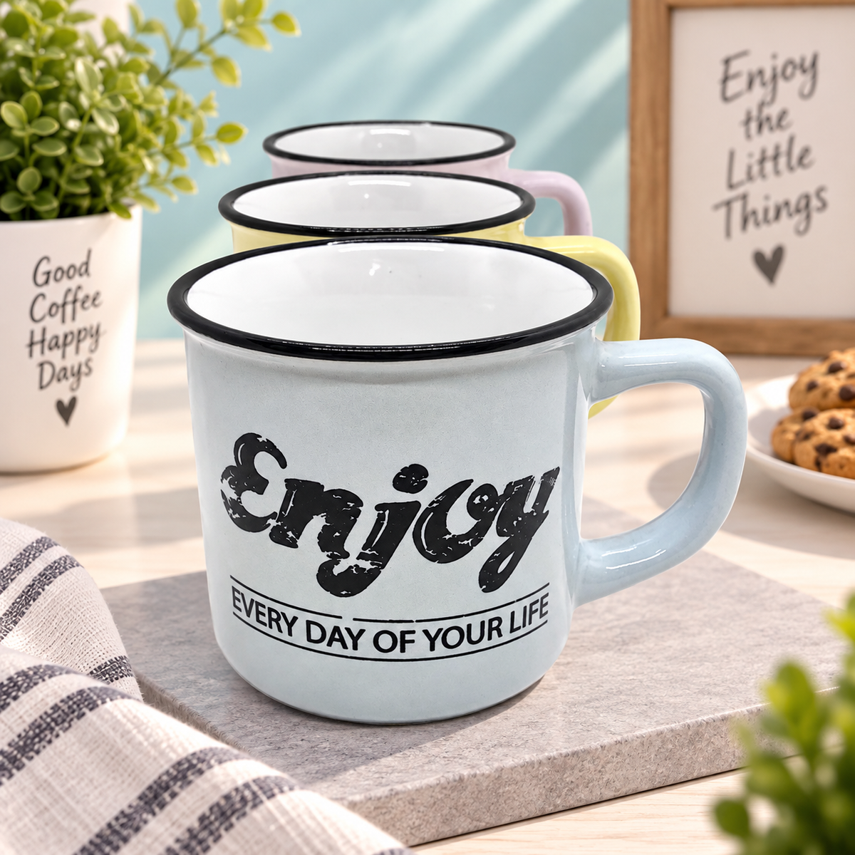 A light blue enamel mug with a black rim and the phrase 'Enjoy EVERY DAY OF YOUR LIFE' printed on the front, styled on a stone tray with two additional pastel mugs behind it in a bright kitchen setting.