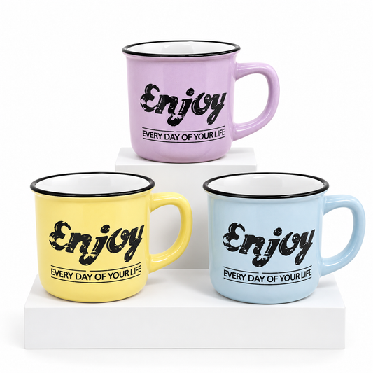 Three enamel-style cups on white display blocks, each printed with the word "Enjoy" and the phrase "EVERY DAY OF YOUR LIFE" in black. The cups are pastel purple, yellow, and light blue with black rims and white interiors.