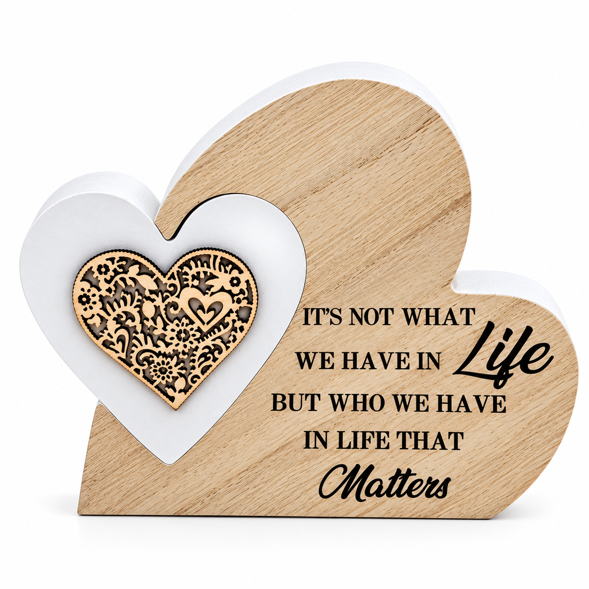A heart-shaped wooden decorative plaque with engraved text reading 'IT'S NOT WHAT WE HAVE IN Life BUT WHO WE HAVE IN LIFE THAT Matters.' A smaller white heart sits in front of the larger natural wood heart, and a laser-cut ornate gold heart is attached on the left side.