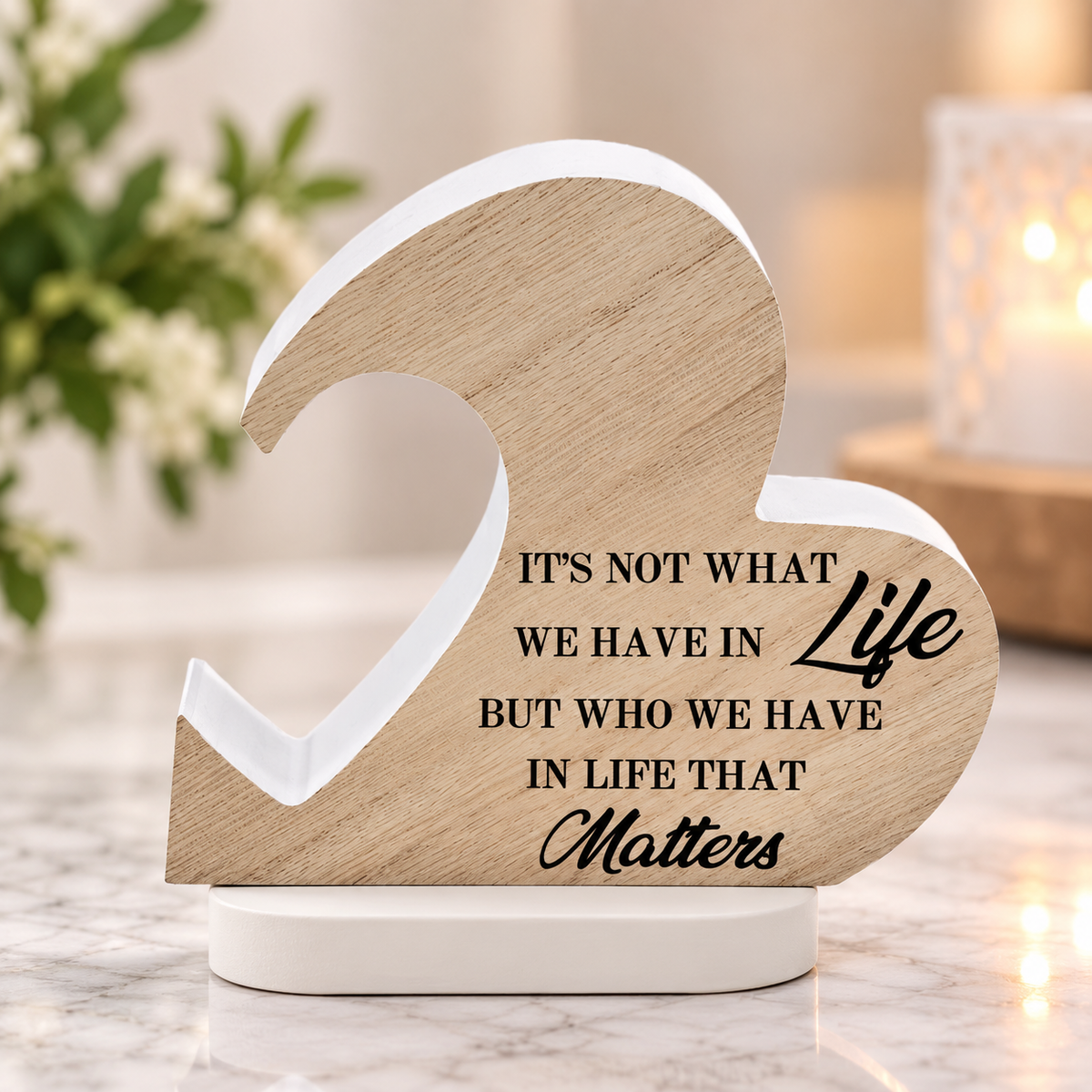 A wooden heart-shaped tabletop sign with a white base, featuring the engraved quote, 'IT'S NOT WHAT WE HAVE IN Life BUT WHO WE HAVE IN LIFE THAT Matters'. The sign is styled on a marble surface with a blurred background containing a lit candle and greenery.