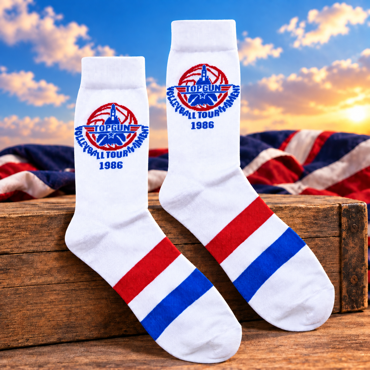 A pair of white crew socks with red and blue stripes and a 'Top Gun Volleyball Tournament 1986' logo, displayed on a wooden crate with a patriotic sky and flag background.