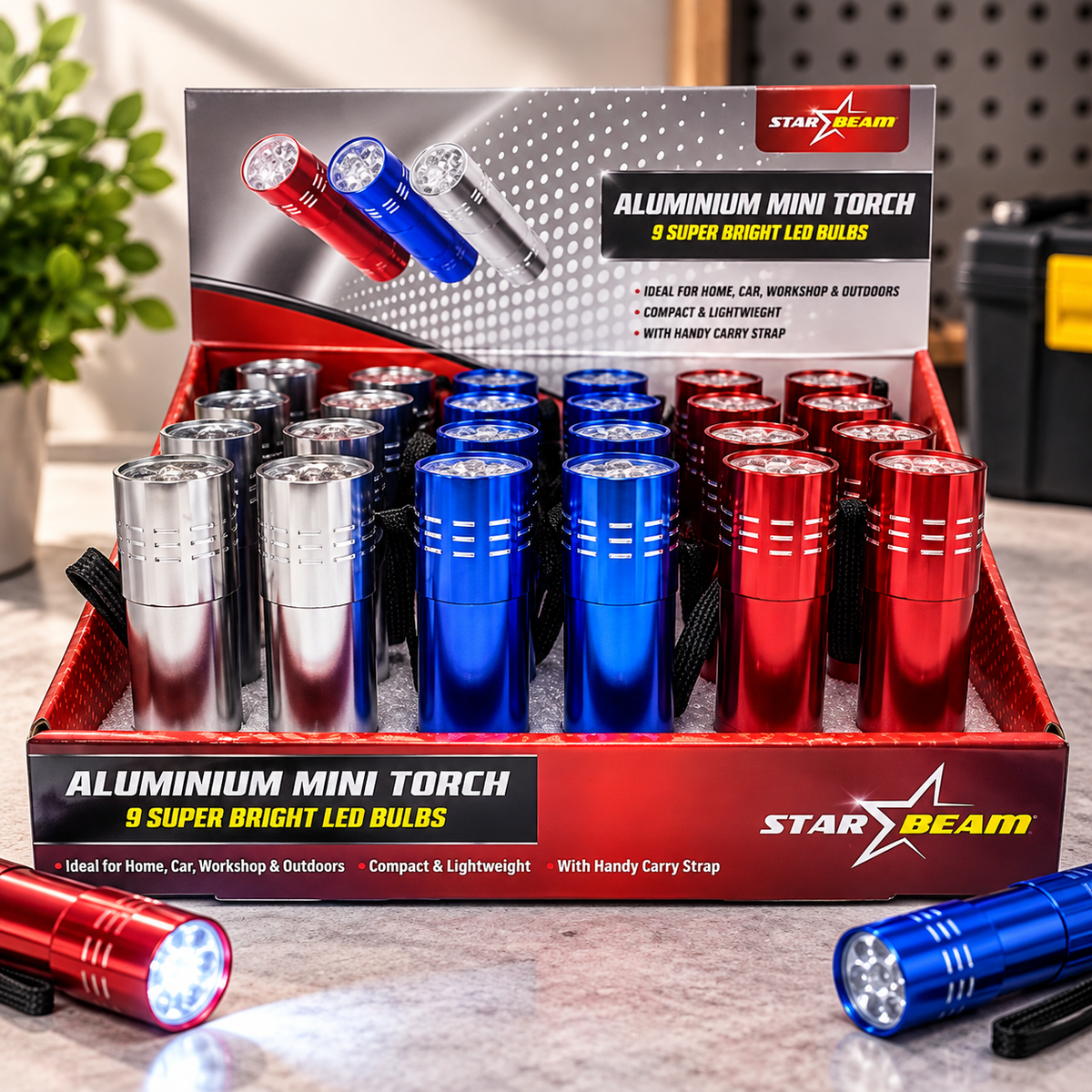 A retail display tray filled with Star Beam aluminium mini torch LED flashlights in red, silver, and blue, with the product box behind them and two flashlights placed in front on a countertop.