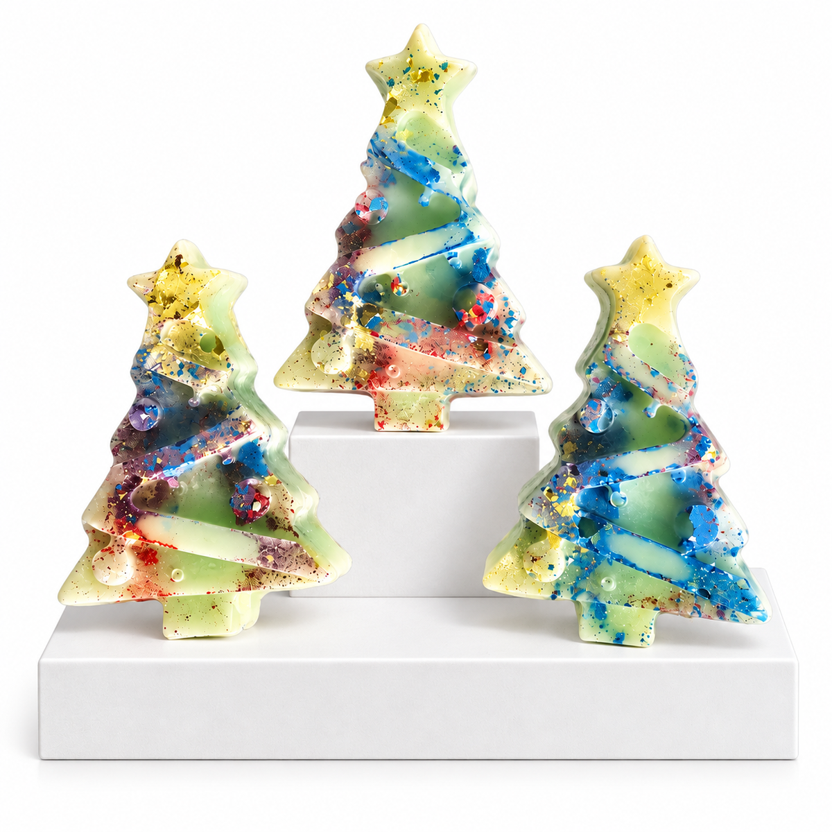 Three small Christmas tree-shaped Wax Melts with star tops, displayed on white risers against a white background. The ornaments have a pale green base with swirling blue, yellow, red, and purple glitter-like patterns.