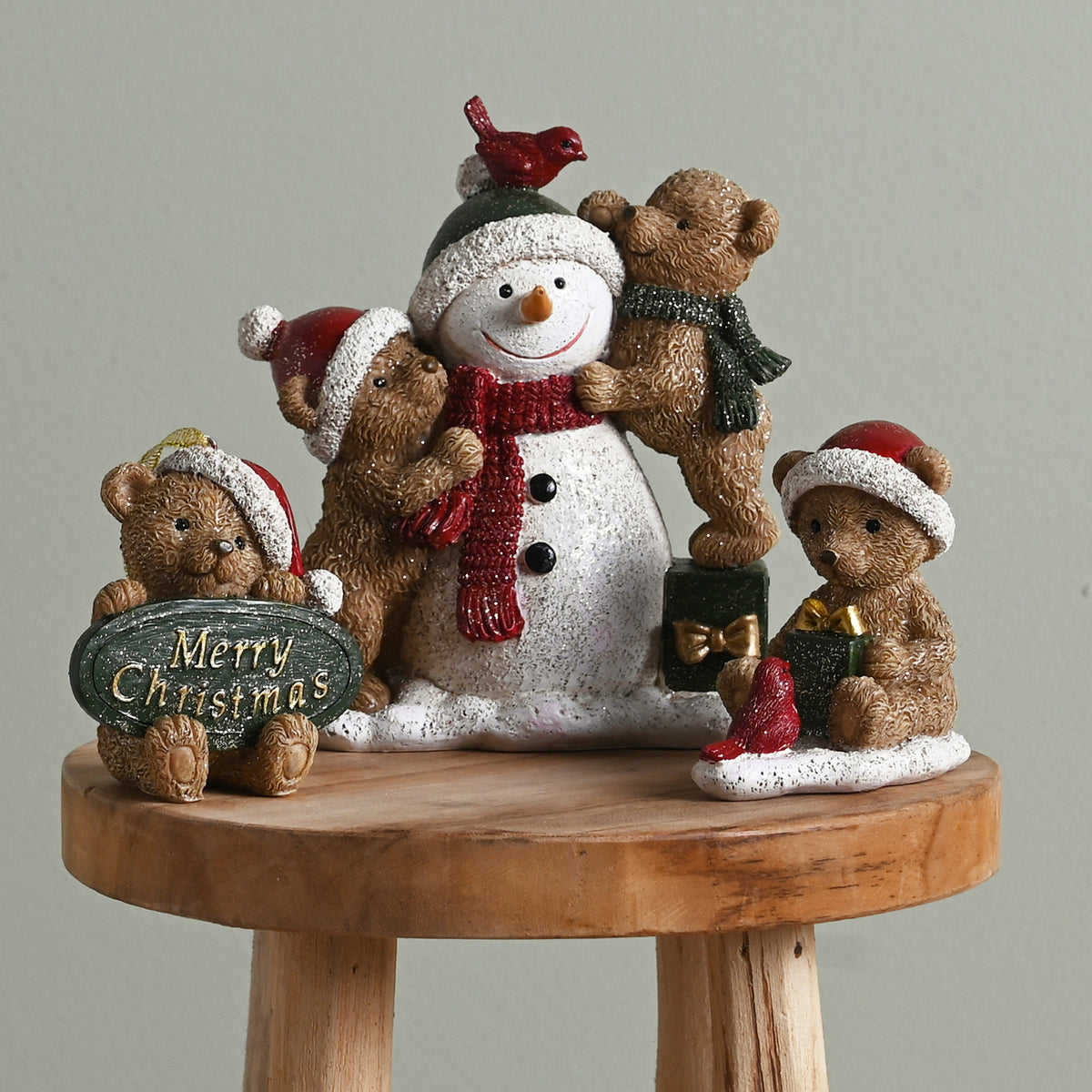 Christmas-themed figurine with snowman and teddy bears on a wooden stool against a gray background