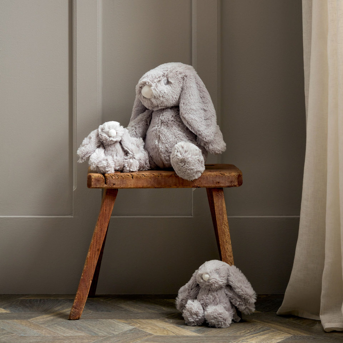 Two gray plush bunny toys on a wooden stool against a neutral wall.