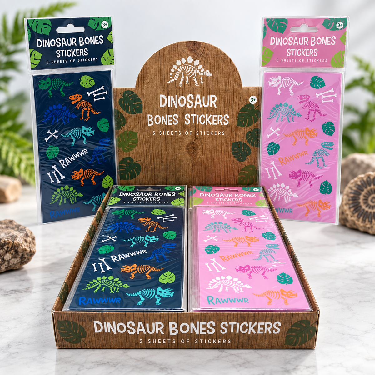 A retail display box filled with dinosaur bones stickers in two colorways: a dark blue pack with green and orange bones, and a pink pack with purple, blue, and orange bones. Two additional sealed packs stand behind the display, one dark blue and one pink.