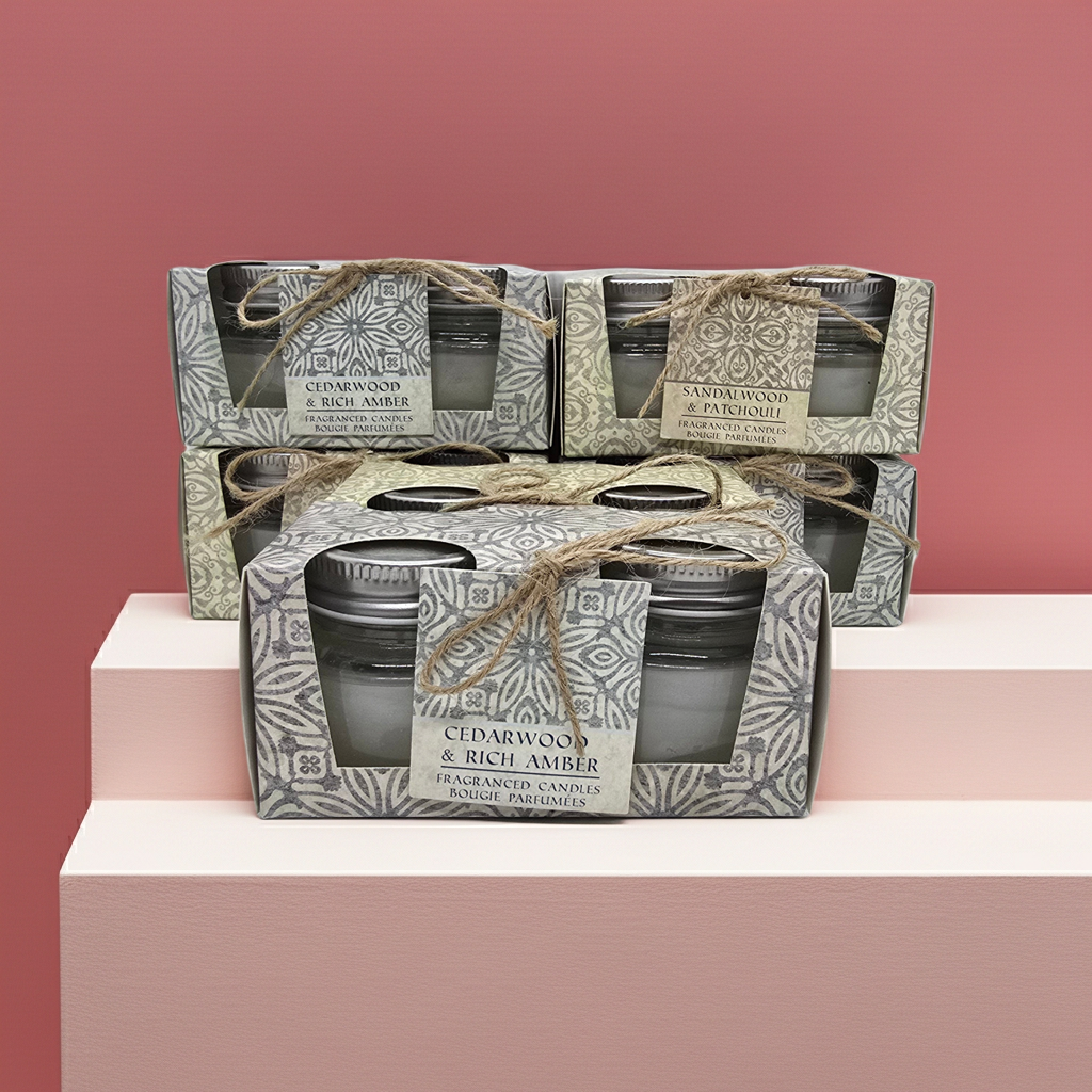 Stack of Scented candle boxes with 2 candles in glass jars per box 1 box has a Cedar wood and rich amber scent and the other has a sandalwood and patchouli scented candles. with decorative packaging on a pink and background.