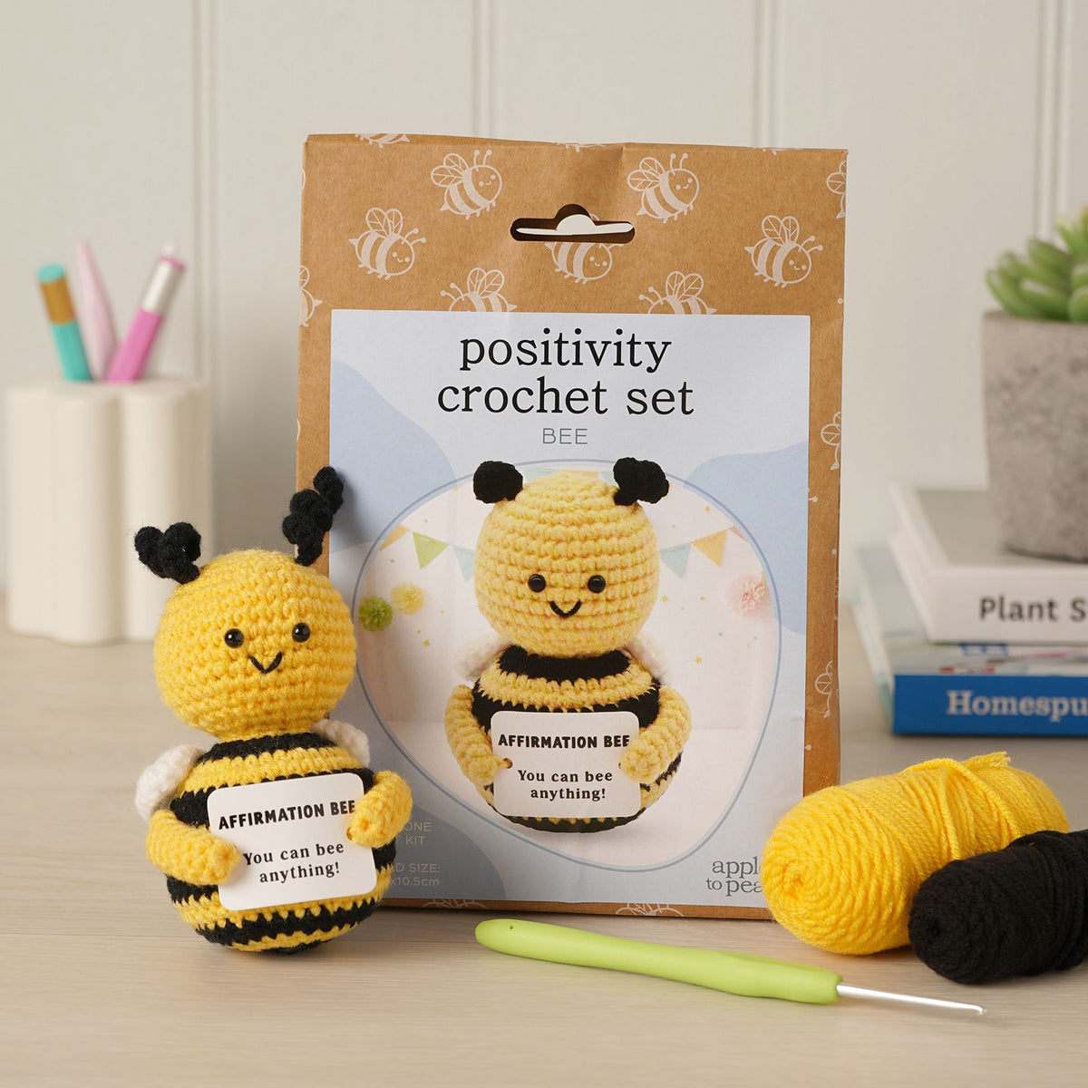 Positivity Crochet Set Bee kit displayed with its kraft paper packaging, a crocheted yellow and black bee affirmation doll, two balls of yarn, and a light green crochet hook on a tabletop.