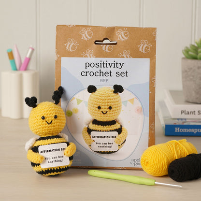 Positivity Crochet Set Bee kit displayed with its kraft paper packaging, a crocheted yellow and black bee affirmation doll, two balls of yarn, and a light green crochet hook on a tabletop.