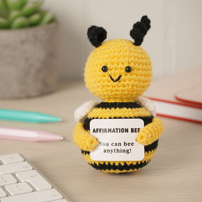 A yellow and black crocheted bee figurine holding a white sign that reads 'AFFIRMATION BEE You can bee anything!' on a light wood desk, with a keyboard, pens, and books blurred in the background.