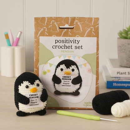 Positivity Crochet Set Penguin kit by Apple to Pen, featuring a finished black and white crocheted penguin with an orange beak in the foreground, its brown pattern packaging box behind it, a green crochet hook, and yarn balls on a tabletop.