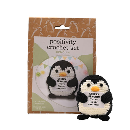 Positivity Crochet Set Penguin, featuring a finished black and white crocheted penguin with a yellow beak in the foreground, next to its brown cardboard packaging box.