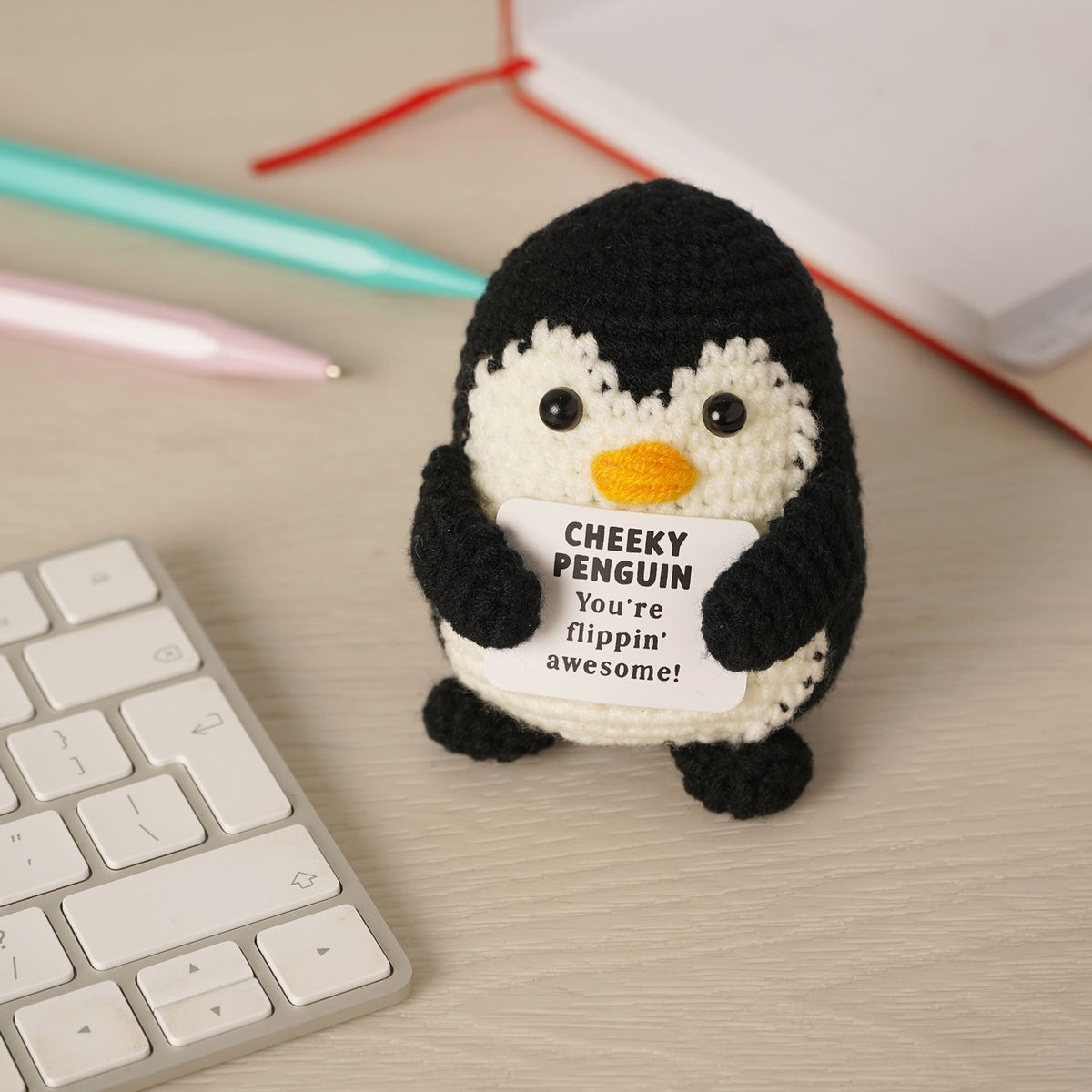 A crocheted penguin toy holding a white sign that reads 'CHEEKY PENGUIN You're flippin' awesome!' sits on a light wood desk next to a keyboard, with pens and a notebook in the background.