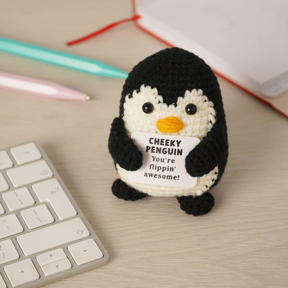 A crocheted penguin toy holding a white sign that reads 'CHEEKY PENGUIN You're flippin' awesome!' sits on a light wood desk next to a keyboard, with pens and a notebook in the background.