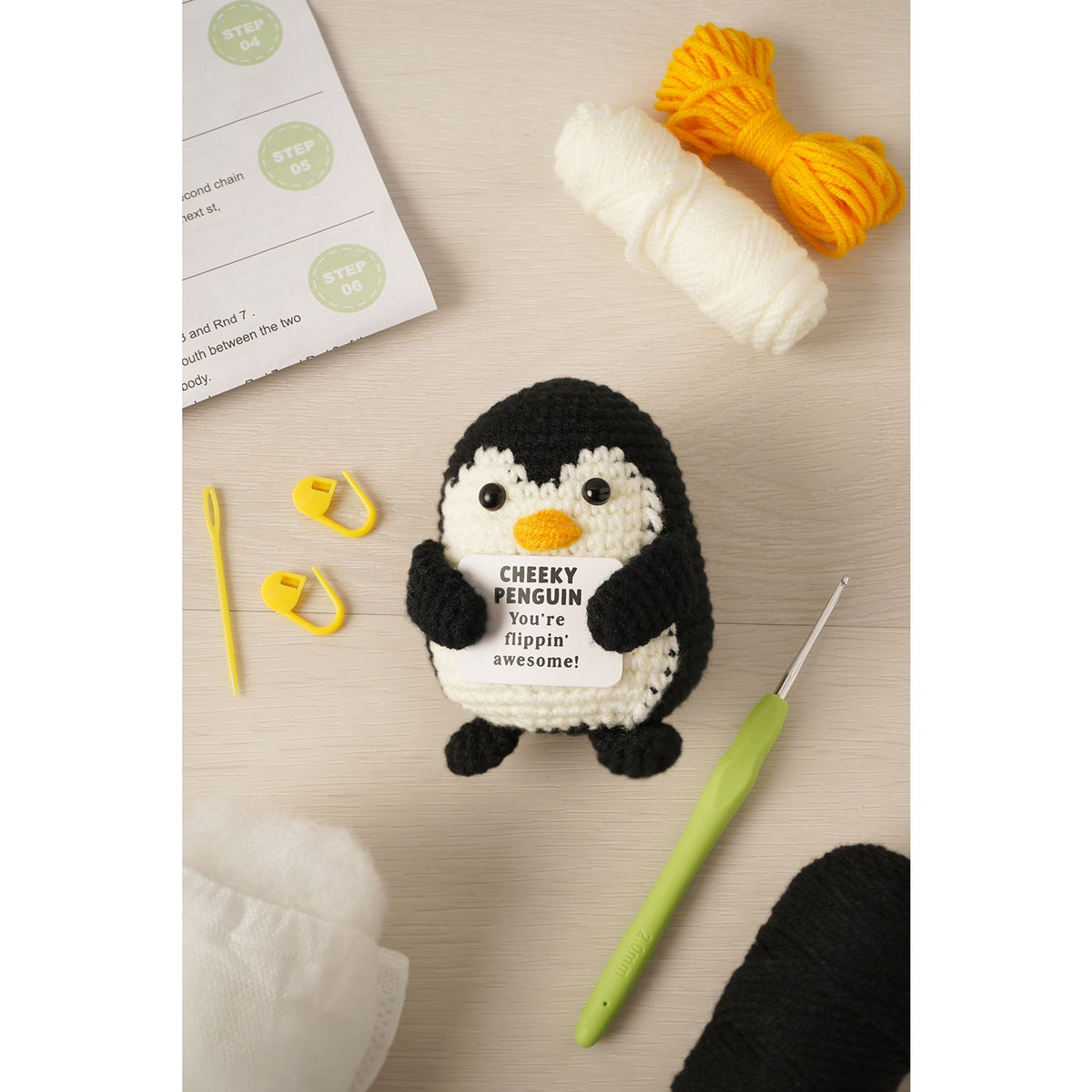 A crocheted penguin toy holding a sign that reads 'CHEEKY PENGUIN You're flippin' awesome!' is arranged on a light wood surface with crochet hooks, yarn, needles, and an instruction sheet around it.