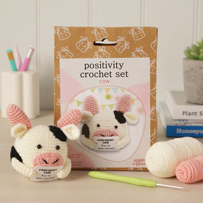 Positivity Crochet Set Cow kit by apple to pear, featuring a crocheted cow toy, a crochet hook, yarn balls, and the product packaging box on a tabletop.