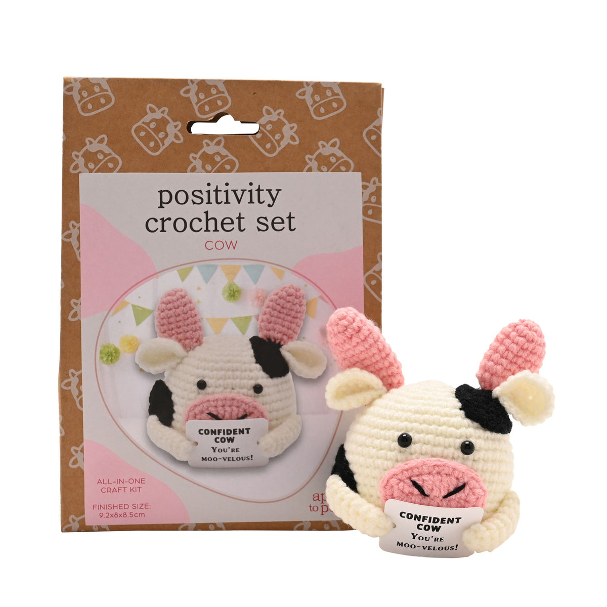 Positivity Crochet Set Cow kit, showing the brown cardboard packaging box alongside the completed crocheted cow toy holding a sign that reads 'CONFIDENT COW YOU'RE MOOD-VALENT!'.