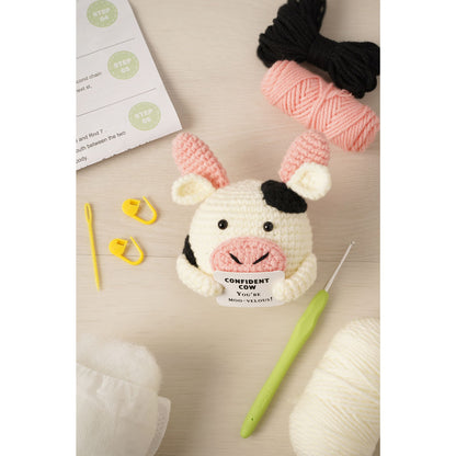 A crocheted cow amigurumi toy holding a sign that reads 'CONFIDENT COW Yoko Mori-Valmont!' is arranged on a light wood surface with yarn balls, a crochet hook, stitch markers, and an instruction sheet.