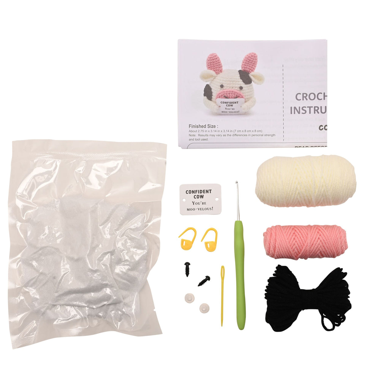 Crochet cow kit with a plastic bag of yarn, a crochet hook, yellow needle holders, a white instruction booklet showing a finished cow, a small card reading 'CONFIDENT CROCHET', and two balls of yarn in cream and pink.