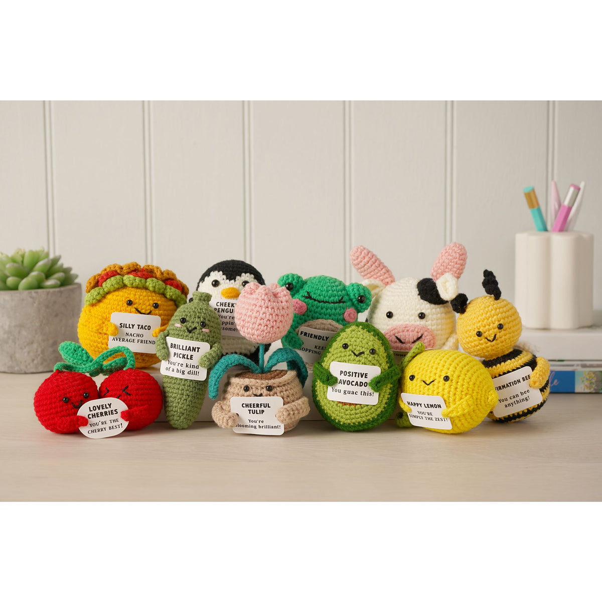 A group of crocheted plush toys shaped like fruit, animals, and characters, arranged on a tabletop in front of a white paneled wall. The toys include a red apple, a yellow citrus fruit, a green avocado, a penguin, a rabbit, a bee, and other small character plushies, each with a small paper tag attached.