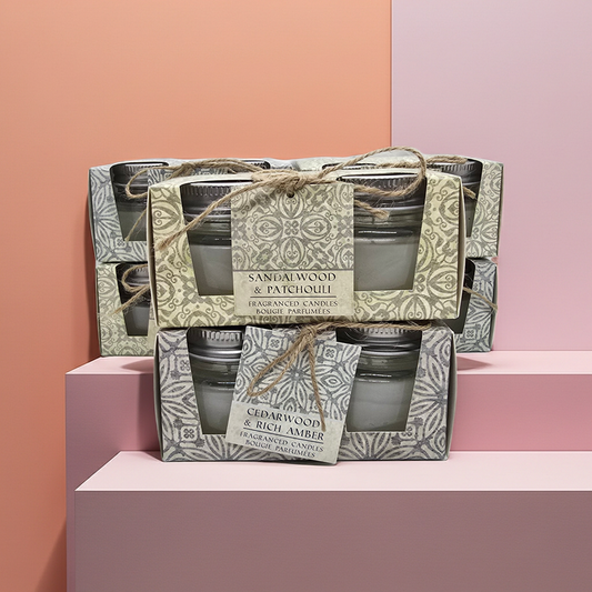 Stack of Scented candle boxes with 2 candles in glass jars per box 1 box has a Cedar wood and rich amber scent and the other has a sandalwood and patchouli scented candles. with decorative packaging on a pink and green gradient background.