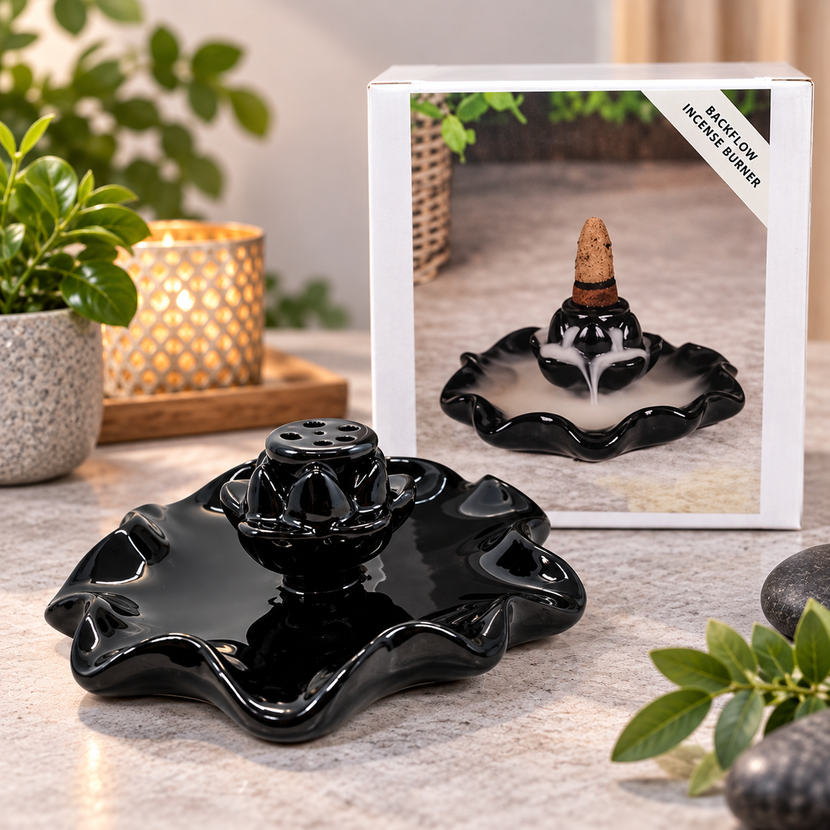 Black ceramic backflow incense burner in the shape of a lotus flower, shown in front of its retail packaging box on a stone countertop with decorative plants and a candle.