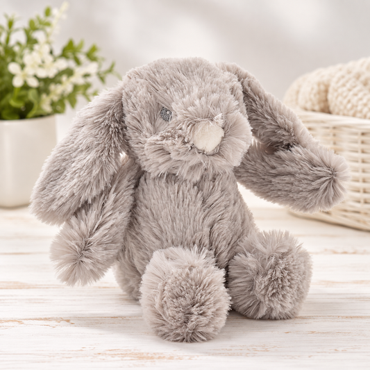 A plush gray bunny stuffed animal with long floppy ears, a soft cream-colored snout, and embroidered eyes, sitting on a light wooden surface with a blurred potted plant and woven basket in the background.