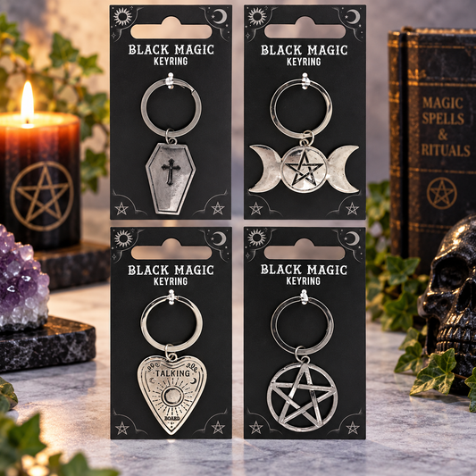 Four black metal keyrings on black retail cards labeled 'BLACK MAGIC KEYRING,' styled on a stone surface with a lit candle, crystal cluster, book, and skull around them.