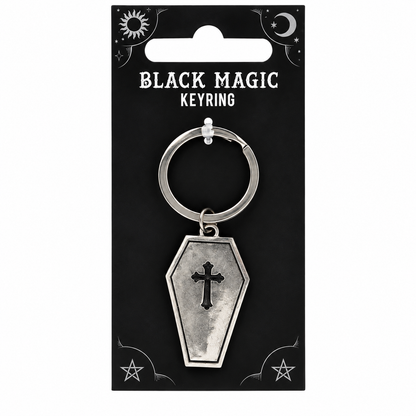A silver coffin-shaped keyring charm with a black cross, attached to a metal key ring and displayed on a black backing card labeled 'BLACK MAGIC KEYRING' with occult-style symbols.