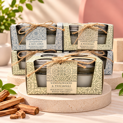 Four packaged scented candles in patterned boxes tied with twine, arranged on a round pedestal with wood bark and greenery in the background. The front box reads 'SANDALWOOD & PATCHOULI FRAGRANCED CANDYBICKS BOUGIE PARFUMÉS'.