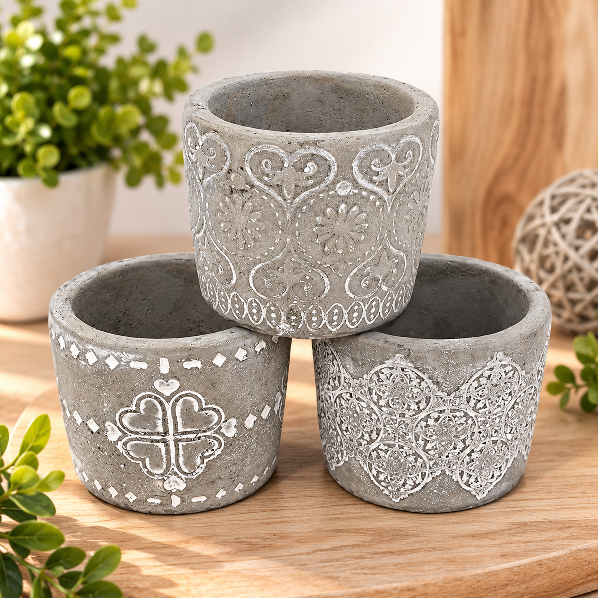 Three small gray decorative planter pots with embossed floral and geometric patterns, arranged in a stacked arrangement on a wooden surface with potted plants in the background.