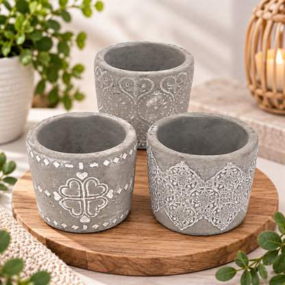 Three gray concrete-style planter pots with white decorative patterns, arranged on a round wooden tray with plants and a warm candle in the background.