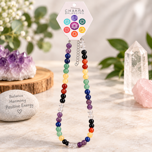 A beaded chakra necklace on a display card, styled with crystals and greenery on a light stone surface. The necklace features multicolor beads representing the chakras, with a silver chain and a small silver heart charm.