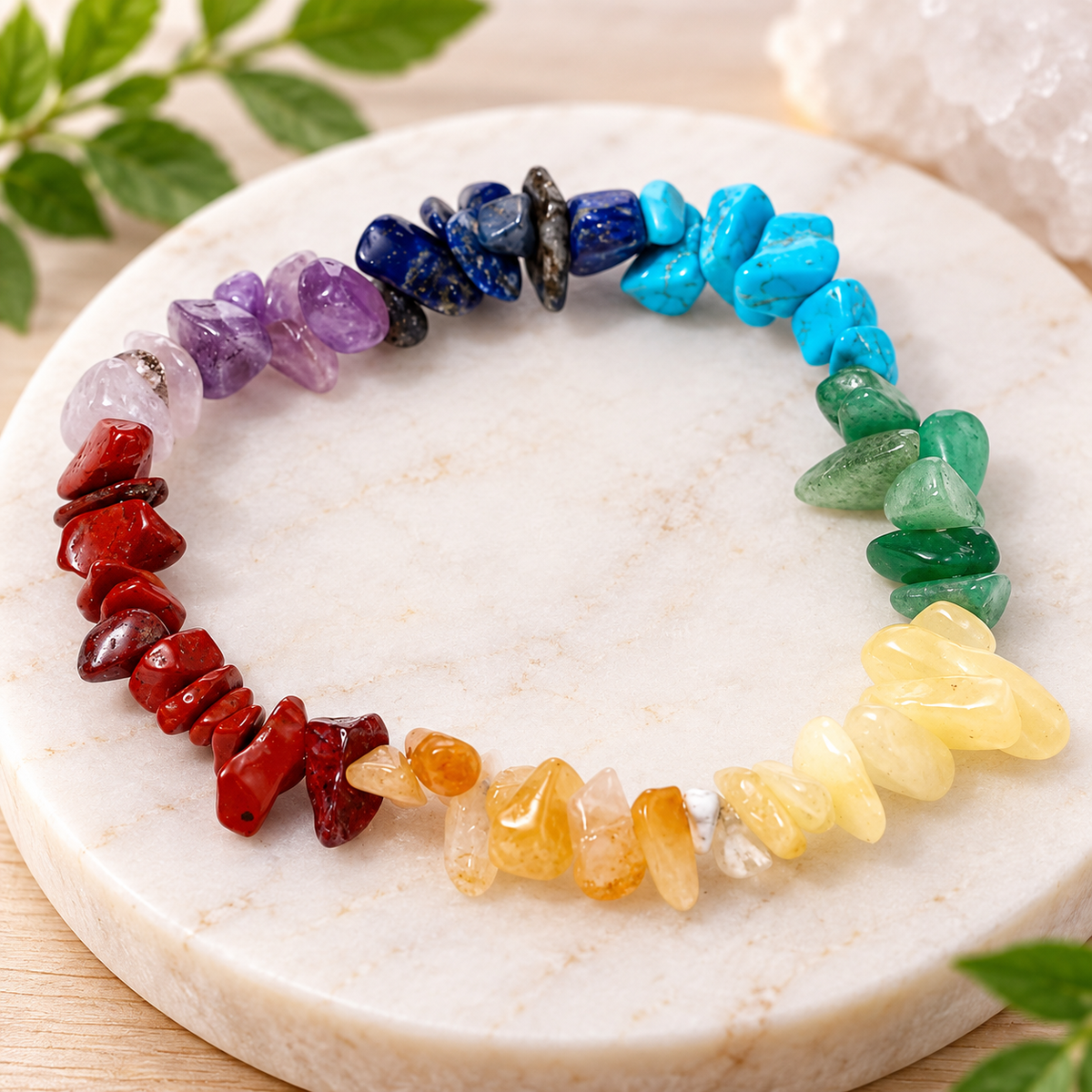 A rainbow crystal chip bracelet arranged in a circle on a round white marble tray, with small green leaves and a crystal cluster in the background.
