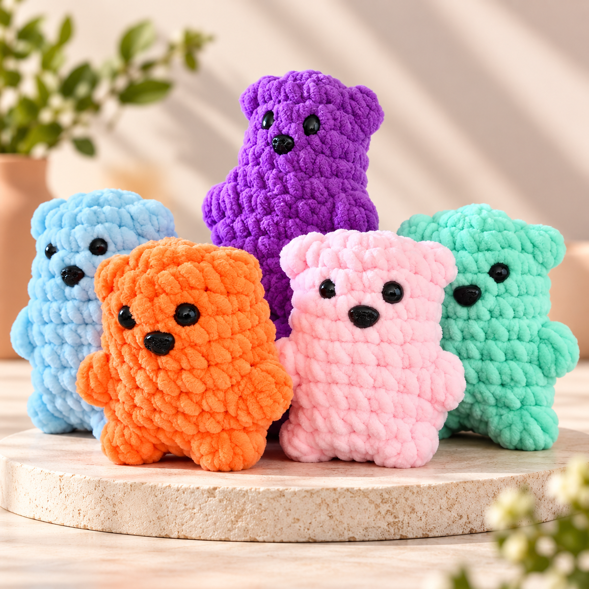Five crocheted plush bear toys in light blue, orange, pink, purple, and mint green, arranged on a round stone pedestal with a softly blurred indoor background.