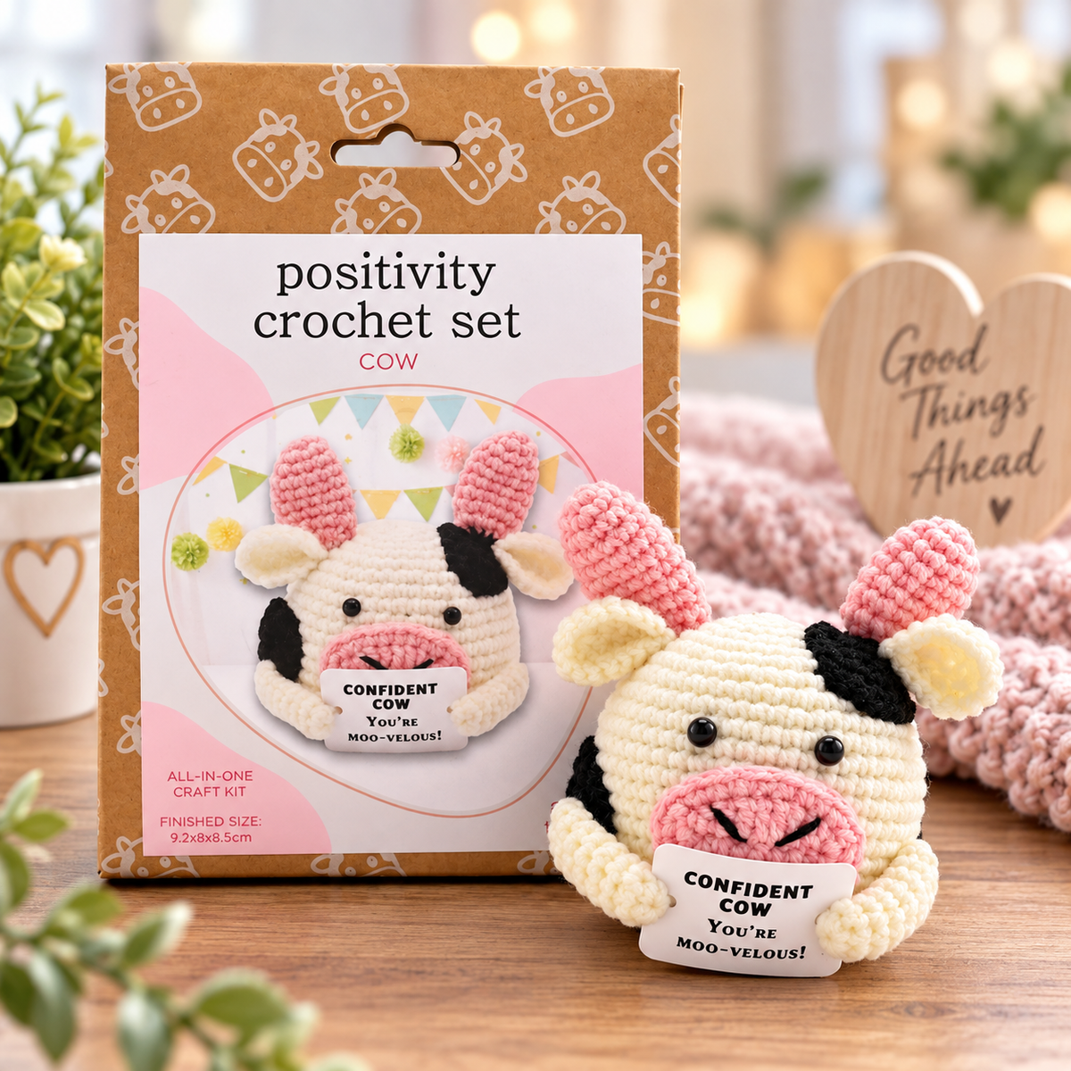 A crocheted cow plush toy sits in front of its retail packaging on a wooden surface. The package reads 'positivity crochet set COW' and shows the cow design on the front.
