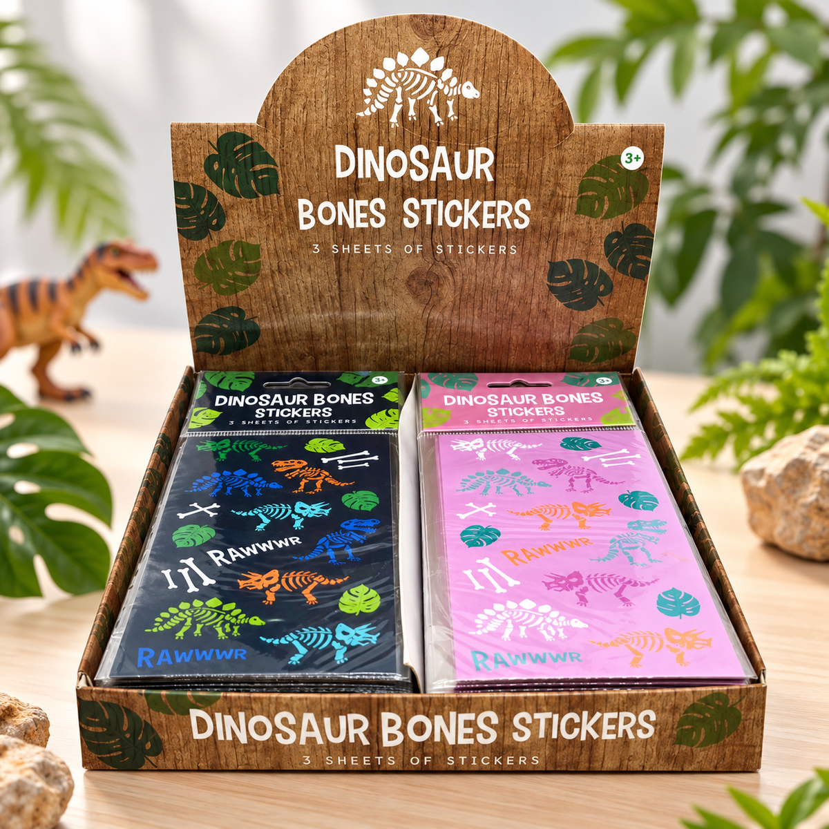 A retail display box filled with two sheets of dinosaur bones stickers, shown in a styled setting with tropical leaves and a small toy dinosaur. The left sheet is dark blue with multicolored bones, and the right sheet is pink with multicolored bones.