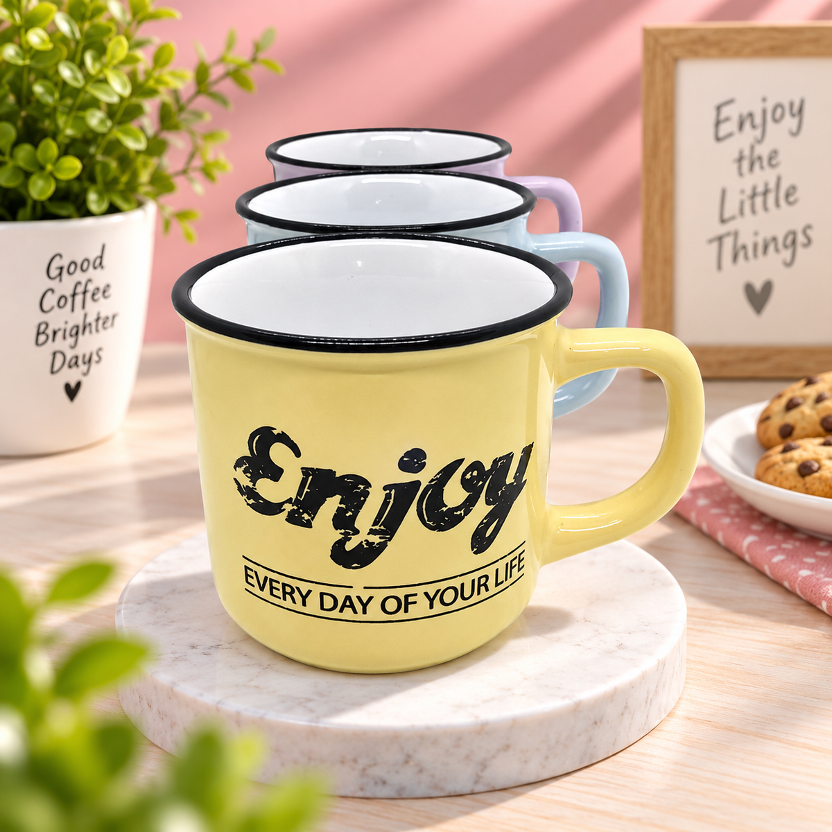 A yellow enamel mug with a black rim and the text 'Enjoy EVERY DAY OF YOUR LIFE' sits on a marble coaster. Three stacked mugs in pastel colors are behind it on a wooden table, with a potted plant, cookies, and a framed sign in the background.