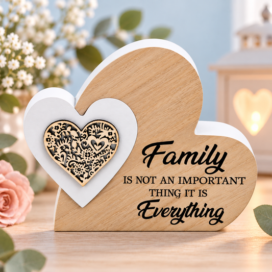 A heart-shaped wooden plaque with black text reading 'Family IS NOT AN IMPORTANT THING IT IS Everything' and a white heart inset featuring a gold floral heart design, displayed on a tabletop with flowers and a lantern in the background.