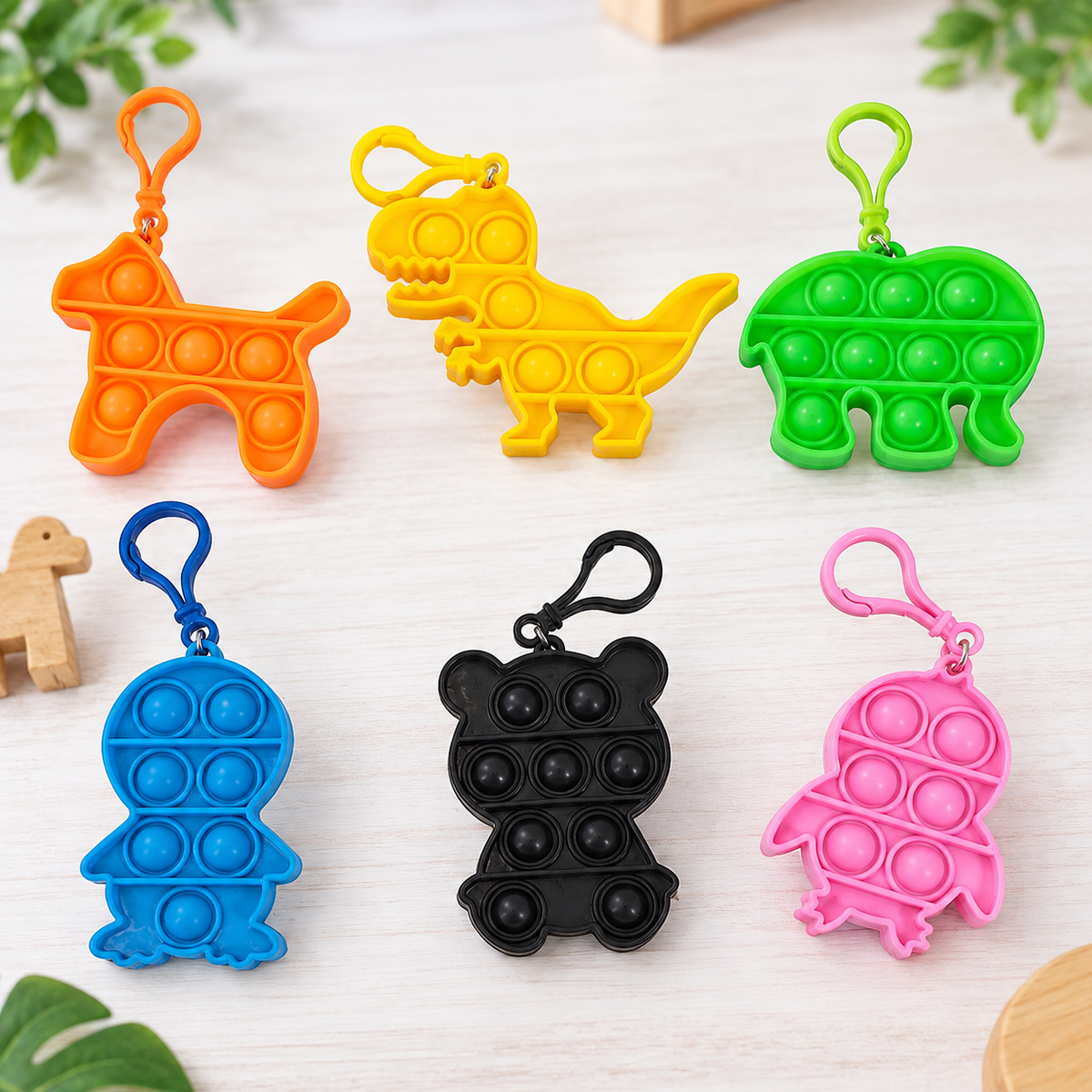 Six colorful fidget pop toy keychains shaped like a dog, dinosaur, elephant, turtle, bear, and chick, arranged on a light wood surface with green leaves in the background.